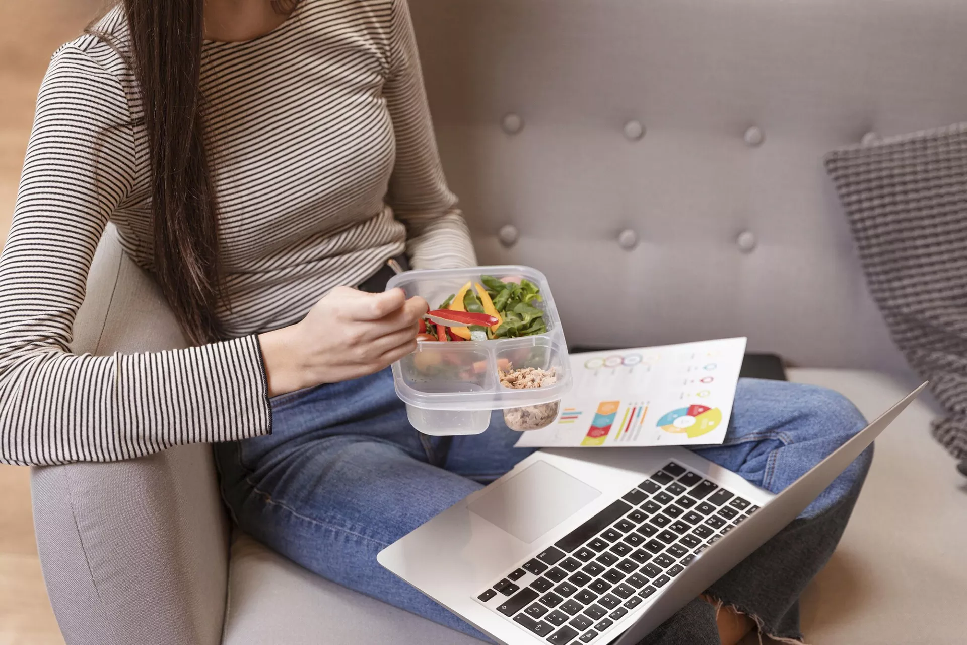 Mujer sentada en un sofá comiendo una ensalada mientras observa un portátil